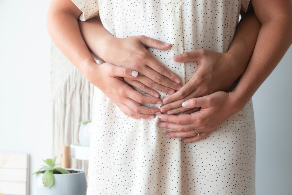 Ein Mann steht hinter einer schwangeren Frau und beide legen ihre Handflächen auf den Babybauch.