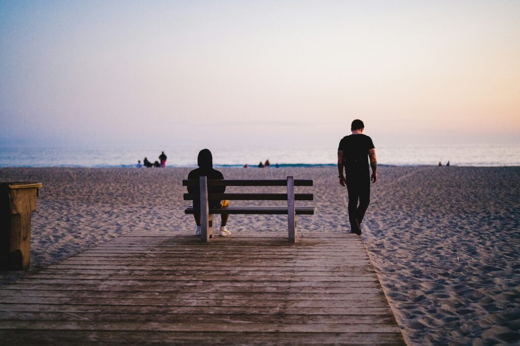 Ein Mann sitzt alleine mit dem Rücken zur Kamera auf einer Bank an einem Strand und schaut auf das Meer hinaus.