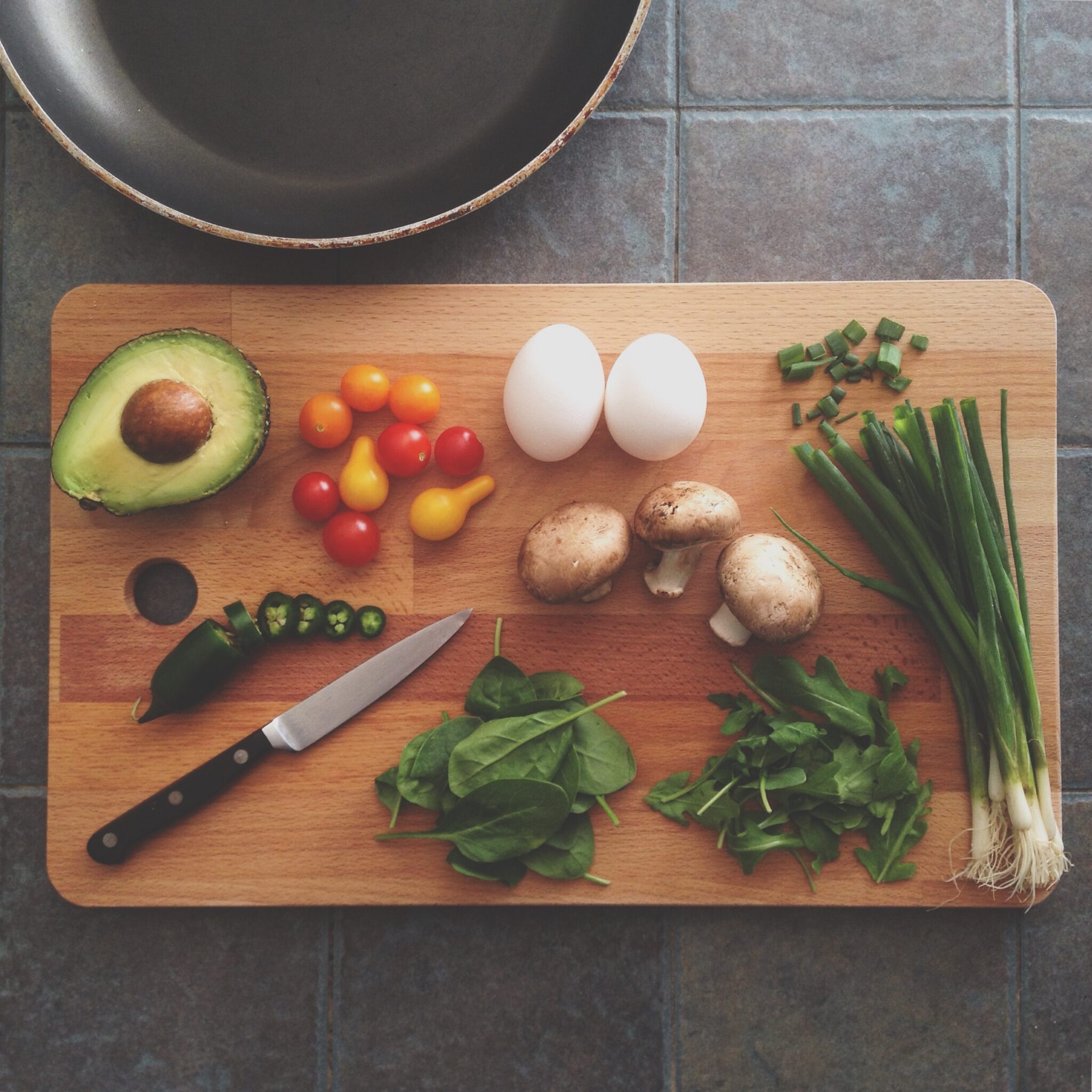 Auf dem Bild ist ein Holzschneidebrett, auf grauem Hintegrund zu sehen. Darauf liegt Gemüse und ein Messer.