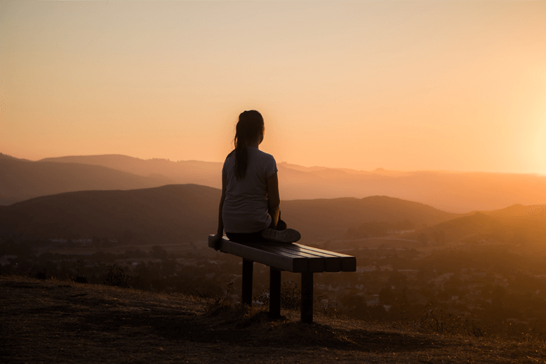 Eine Frau sitzt während eines Sonnenuntergangs auf einer Bank auf einem Berg mit Blick ins Tal.