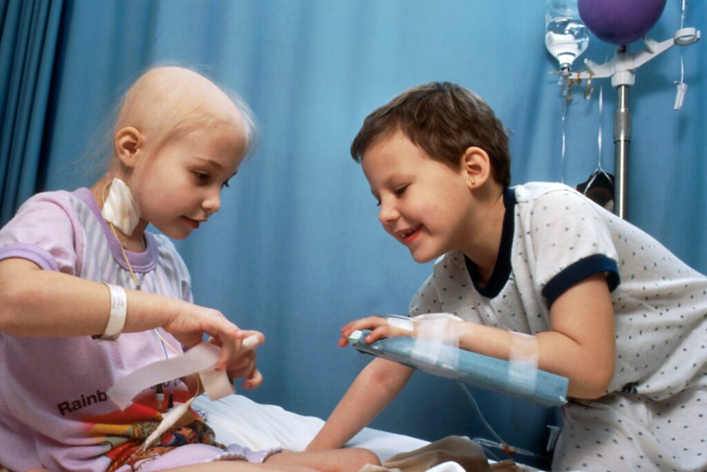 Zwei Kinder spielen im Krankenhaus auf einem Bett. Beide sehen schwer krank, aber in diesem Moment glücklich aus und lachen.