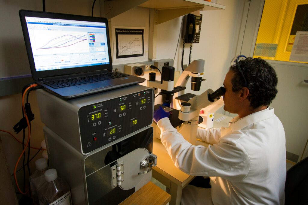 Ein Wissenschaftler schaut durch ein Mikroskop Proben an. Auf einem Bildschirm neben ihm wird eine Grafik angezeigt.