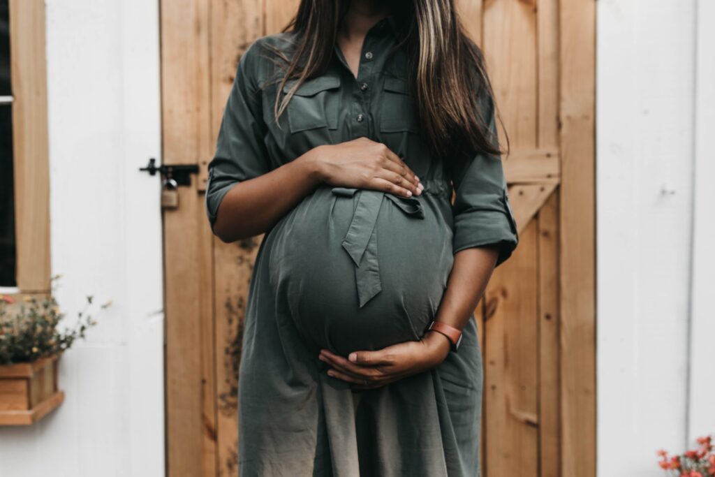 Eine schwangere Frau in einem grünen Kleid umfasst ihren Babybauch mit beiden Händen.